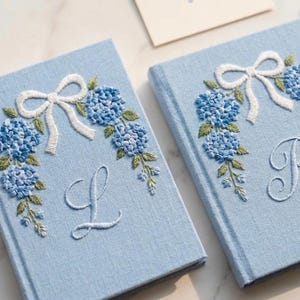 May include: Two light blue notebooks with embroidered floral designs. Each notebook features a white bow, blue hydrangea blooms, green leaves, and a single initial. The notebooks are closed and appear to be made of fabric.