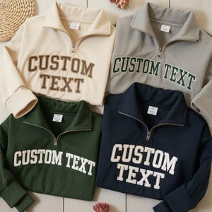 May include: Four quarter-zip sweatshirts in cream, gray, dark green, and navy blue. Each sweatshirt has the text "CUSTOM TEXT" in a contrasting color. The cream sweatshirt has brown text, the gray has green, the dark green has white, and the navy blue has white.