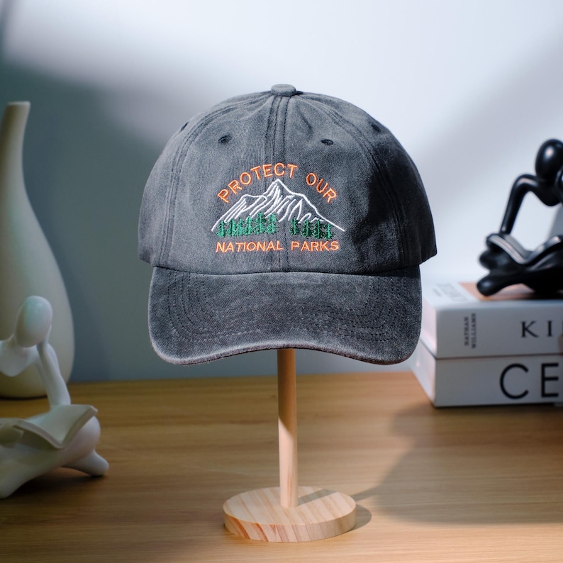 May include: A grey baseball cap with the text "PROTECT OUR NATIONAL PARKS" embroidered in orange and green. The cap features a mountain and tree design. The hat is displayed on a wooden hat stand.