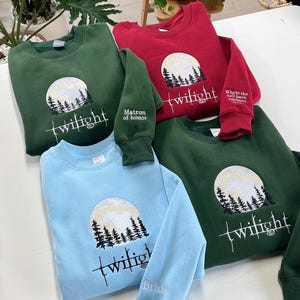 May include: A collection of sweatshirts in various colors, including green, red, and blue. Each sweatshirt features an embroidered design of a full moon, trees, and the word "twilight." Some sleeves have additional text, such as "Matron of honor" and "Bride."