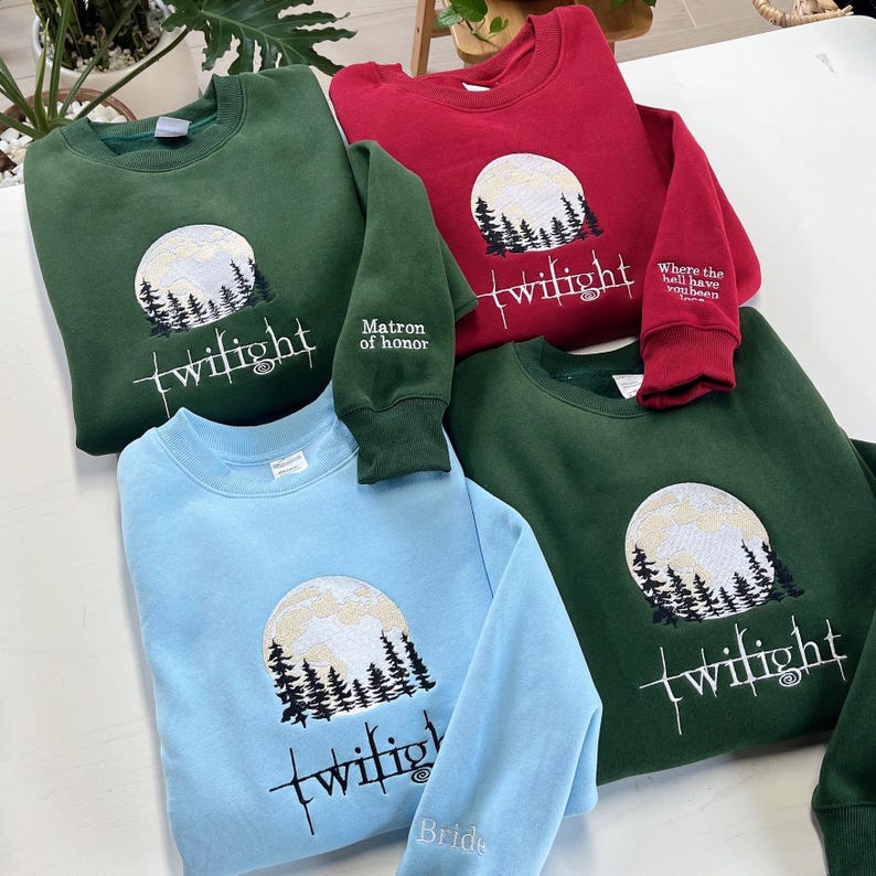 May include: A collection of sweatshirts in various colors, including green, red, and blue. Each sweatshirt features an embroidered design of a full moon, trees, and the word "twilight." Some sleeves have additional text, such as "Matron of honor" and "Bride."