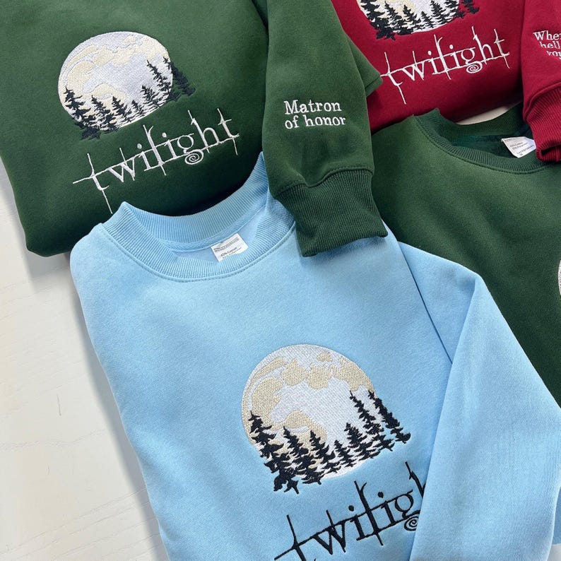 May include: Crewneck sweatshirts in various colors, including green, light blue, and burgundy. Each sweatshirt features an embroidered design of a full moon, trees, and the word "twilight". One sleeve reads "Matron of honor".
