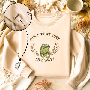 May include: Cream-colored sweatshirt featuring a green frog illustration with the text "AIN'T THAT JUST THE WAY?" and a floral wreath. The left sleeve has a coffee cup graphic and the word "Custom" on the cuff.