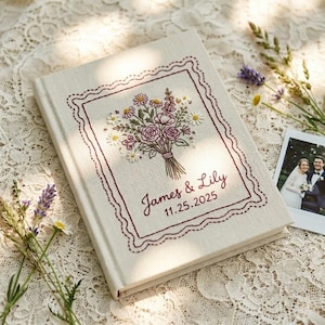May include: A cream-colored fabric-covered book featuring an embroidered bouquet of flowers in shades of pink, green, and white, enclosed in a dark red scalloped border. The text James & Lily and the date 11.25.2025 are embroidered in dark red.