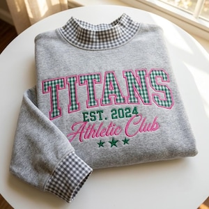 May include: A heather gray sweatshirt featuring a mock neck collar and sleeve cuffs in a gray and white gingham pattern. The front displays the embroidered text TITANS in green gingham with pink outlines, EST. 2024, and Athletic Club in pink.