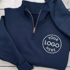 May include: Navy blue quarter-zip sweatshirt with a silver zipper. The sweatshirt features a white embroidered logo that says "YOUR LOGO HERE" in a circular design. The fabric appears to be a soft, comfortable material.