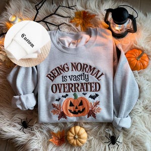 May include: A light gray sweatshirt with the embroidered phrase "Being Normal is Vastly Overrated" above a jack-o'-lantern and bats. The sleeve cuff has the word "Custom" embroidered on it. The image has Halloween decorations.