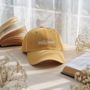 May include: Mustard yellow baseball cap with the embroidered text "well-read" in white. The cap is sitting on a wooden surface next to an open book. The background includes a sheer white curtain and dried flowers.