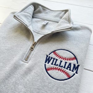 May include: A light gray sweatshirt with a quarter-zip collar. The sweatshirt features a baseball design with the name "WILLIAM" embroidered in navy blue and red thread. The baseball is inside a navy blue circle.