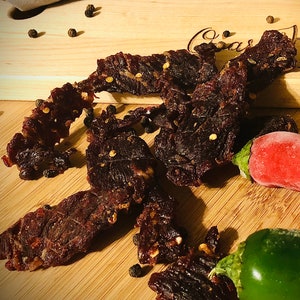 May include: Close-up of beef jerky with peppercorns and red pepper flakes on a wooden cutting board. A red chili pepper, green chili pepper, and lemon are also visible. The cutting board has the text "Oscar's Kitchen 2015-2020".