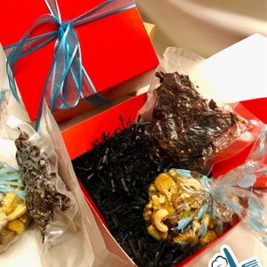 May include: A red gift box with a blue ribbon and bow, filled with a clear bag of dried beef jerky and a clear bag of nuts. The box is sitting on a white surface and has a black and white patterned paper inside. The box is labeled " Opa's Kitchen".