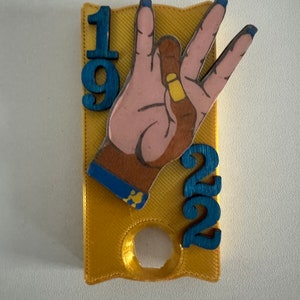 Puede incluir: Una placa de color dorado impresa en 3D con los números 19 y 22 a cada lado. Una mano de madera con uñas azules está en el centro de la placa, haciendo el gesto de la mano 'W'.