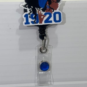 May include: A blue and white badge holder with a cartoon image of a woman with a blue headscarf and the text "1920". The badge holder has a blue gem on the bottom.