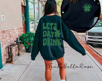 Puede incluir: Una sudadera negra con el texto "Let's Day Drink" y un trébol verde. La sudadera la lleva puesta una persona que está de pie en una acera frente a un edificio.