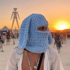 May include: A light blue, chunky knit balaclava featuring long, floppy rabbit-style ears. The headgear is crafted from a soft, plush yarn with a visible, thick looped texture that covers the head and neck, leaving an opening for the eyes.