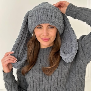 May include: A dark gray beanie made of thick, chunky yarn featuring two long, dangling rabbit-style ears on the sides. The hat has a ribbed texture and is crafted from a soft, plush material that covers the top of the head and hangs down.