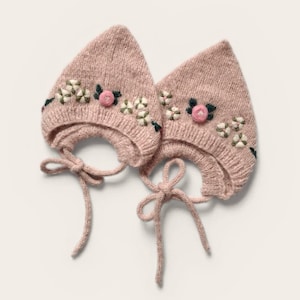 May include: Two dusty pink pointed bonnets made of knit fabric, each featuring embroidered floral details with small white petals and a central pink rosebud. Both bonnets include matching knit chin ties for securing the headwear.