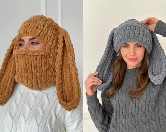 May include: Two chunky knit balaclavas made from soft, thick yarn. One is a solid tan color featuring long, drooping rabbit-style ears and a face-covering design. The other is a solid gray color, styled as a beanie with long, hanging rabbit ears.