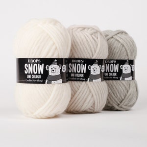 May include: Three skeins of Drops Snow yarn in white, beige, and light grey. Each skein features a black paper label with the text Drops Snow Uni Colour, a graphic of a bear wearing a hat and scarf, and the text 100% Wool and Excellent for felting.