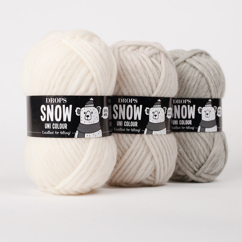 May include: Three skeins of Drops Snow yarn in white, beige, and light grey. Each skein features a black paper label with the text Drops Snow Uni Colour, a graphic of a bear wearing a hat and scarf, and the text 100% Wool and Excellent for felting.
