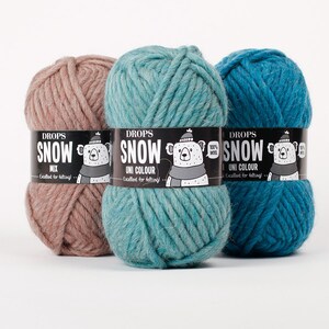May include: Three skeins of thick yarn in dusty pink, light teal, and dark blue. Each skein features a black paper label with the text DROPS SNOW, a graphic of a bear wearing a hat and scarf, and the phrase Excellent for felting.