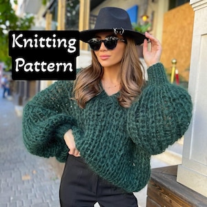 May include: A green knitted sweater with a V-neck and long sleeves. The sweater has a loose, oversized fit and a textured knit pattern. The model is wearing the sweater with black pants and a black hat.