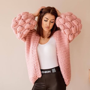 Puede incluir: Un cárdigan de punto grueso rosa claro con mangas texturizadas y de gran tamaño. El suéter abierto está hecho de un hilo grueso y tiene un ajuste relajado. La modelo lleva una camiseta blanca y leggings negros.