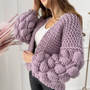 Op de afbeelding: Een lila gebreide cardigan met een grove breistructuur en grote, bolle mouwen. De cardigan is open aan de voorkant en heeft een V-hals.