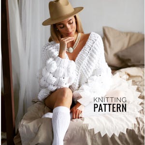 Puede incluir: Una mujer con un sombrero fedora beige, un suéter blanco de punto grueso y calcetines blancos hasta la rodilla. El suéter tiene un escote en V profundo y mangas abullonadas. El texto "KNITTING PATTERN" es visible.
