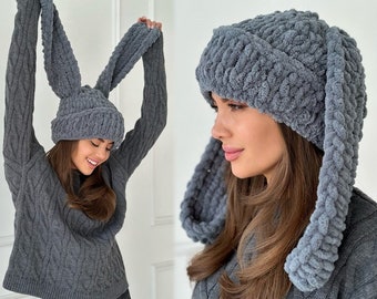 May include: A dark gray beanie made of thick, plush, chunky yarn featuring two long, dangling rabbit-style ears. The hat has a ribbed cuff and a soft, textured knit construction that provides a voluminous appearance and a cozy, tactile finish.