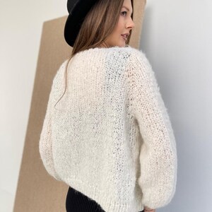 May include: A cream-coloured, chunky knit jumper with long sleeves and a loose, textured weave. The jumper features a wide neckline and a slightly cropped hem. A solid black fedora hat with a wide brim is worn above the jumper.