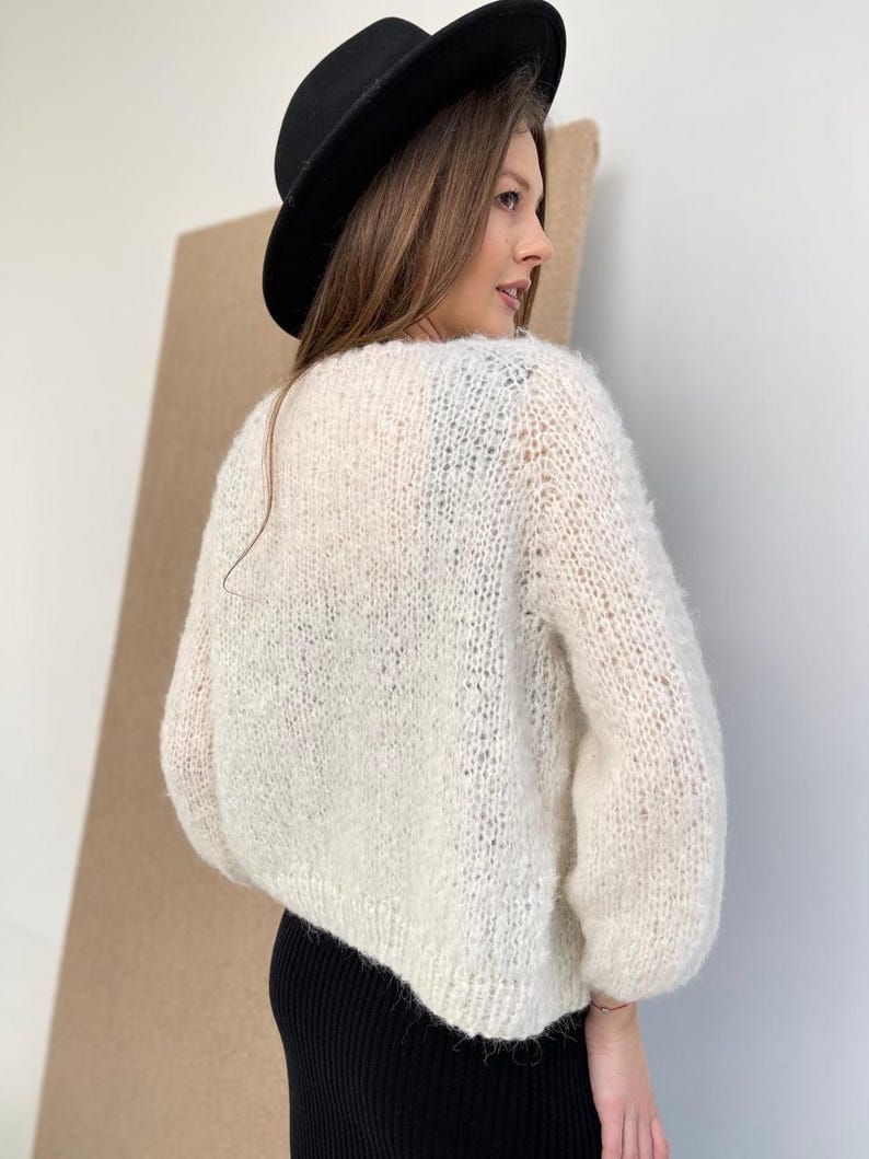 May include: A cream-coloured, chunky knit jumper with long sleeves and a loose, textured weave. The jumper features a wide neckline and a slightly cropped hem. A solid black fedora hat with a wide brim is worn above the jumper.