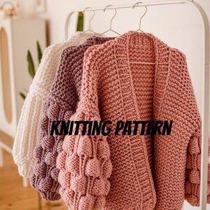 Puede incluir: Tres cárdigans de punto con mangas abullonadas en blanco, morado y rosa. Los cárdigans están colgados en un perchero blanco. El texto "KNITTING PATTERN" es visible en la imagen.