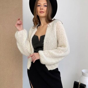 May include: A black wide-brimmed hat, a cream-coloured fuzzy knit cardigan with long sleeves, a black sleeveless top with a notched neckline, and a black ribbed midi skirt featuring a side slit. The outfit is styled with a small black handbag.