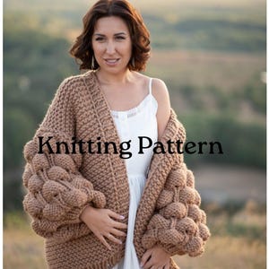 Puede incluir: Un cárdigan de punto grueso color canela con mangas extragrandes que presentan una textura de puntos de burbuja. La prenda se lleva sobre un vestido blanco de tirantes. El texto Knitting Pattern aparece superpuesto en negro.