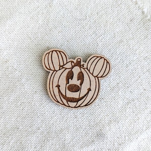 May include: A wooden pumpkin carving with a Mickey Mouse face. The pumpkin has a stem and a carved smile. The carving is a light brown colour.