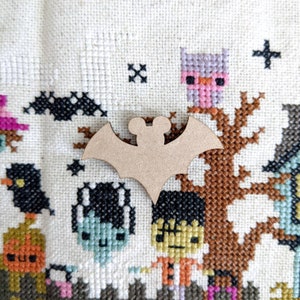May include: A brown wooden bat cutout on a white background with a cross-stitch pattern featuring various Halloween characters, including an owl, a ghost, and a Frankenstein.