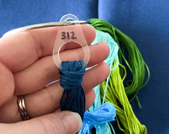 May include: A close-up of a hand holding a blue embroidery floss with a plastic thread organizer labeled "312". The floss is surrounded by other embroidery floss in various shades of blue and green.