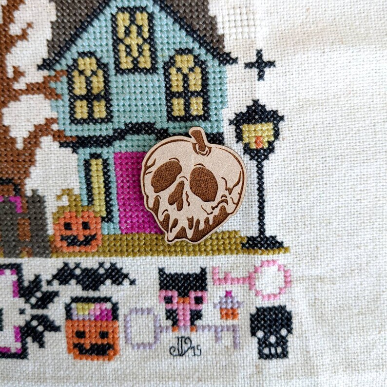 May include: A wooden, brown, hand-painted, poison apple with a skull design. The apple is dripping with a white substance. The apple is on a white background with a cross-stitch pattern.