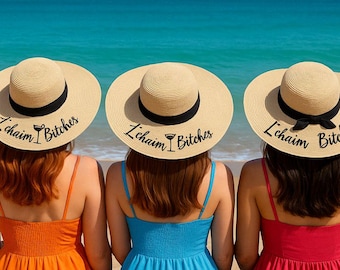 Pode incluir: Três mulheres sentadas numa praia arenosa usam chapéus de sol de palha com faixas pretas e o texto "L'chaim Bitches". Elas usam vestidos laranja, azuis e vermelhos. Os chapéus têm um gráfico de copo de coquetel.