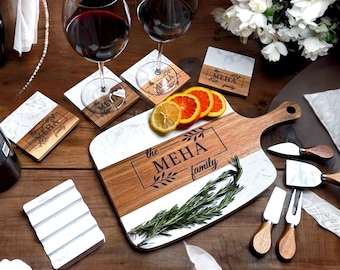 May include: A personalized wooden and marble serving board with the text "the MEHA family" engraved on it. The board is surrounded by four coasters with the same text and a small white dish with four grooves. There are also four cheese knives and a sprig of rosemary on the board.