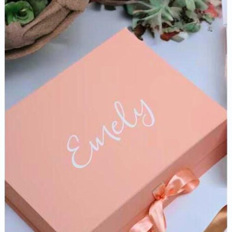 May include: A coral-colored gift box with a matching satin ribbon. The box features the name "Emely" in elegant white script on the lid. The box is closed and ready to be gifted.