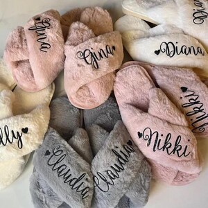 May include: A collection of personalized plush slippers in various colors, including pink, white, and gray. Each slipper features a name embroidered in black script. The names include "Gina", "Diana", "Nikki", "Kelly", and "Claudia".