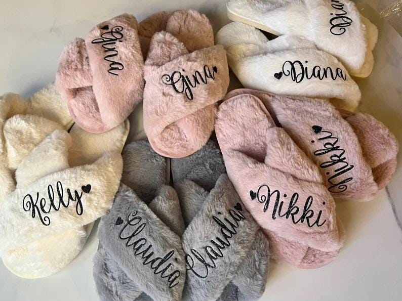 May include: A collection of personalized plush slippers in various colors, including pink, white, and gray. Each slipper features a name embroidered in black script. The names include "Gina", "Diana", "Nikki", "Kelly", and "Claudia".