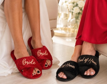 May include: A pair of red plush slippers with the word Bride embroidered in gold script and a pair of black plush slippers with the word Maid of Honor embroidered in gold script, both featuring a crisscross strap design and open toes.