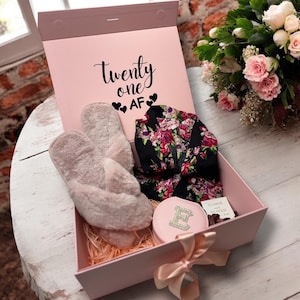 May include: A pink gift box with the text "Twenty one AF" contains a pair of fuzzy slippers, a floral robe, and a pink compact mirror. The box is tied with a satin ribbon and sits on a white wooden surface.