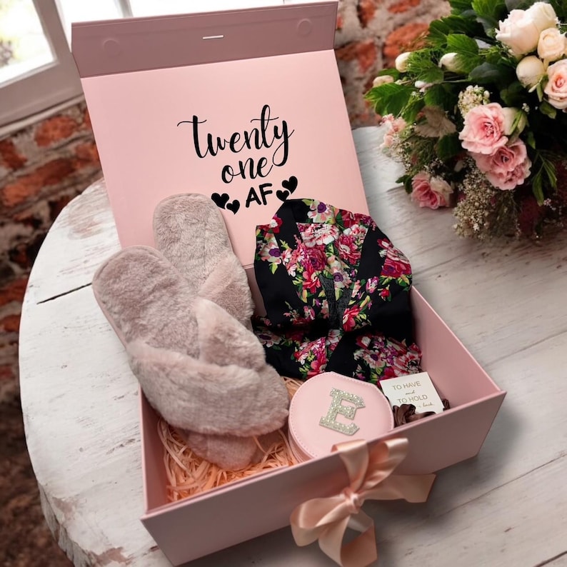 May include: A pink gift box with the text "Twenty one AF" contains a pair of fuzzy slippers, a floral robe, and a pink compact mirror. The box is tied with a satin ribbon and sits on a white wooden surface.