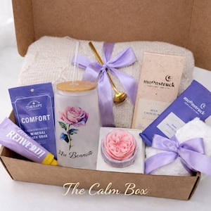 May include: A gift box filled with self-care items. Includes a glass with a rose design, bath products, chocolate bars, a small towel, and a decorative soap. The box is tied with a purple ribbon and contains a white blanket.