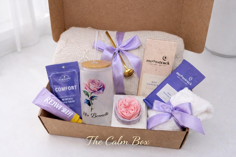 May include: A gift box filled with self-care items. Includes a glass with a rose design, bath products, chocolate bars, a small towel, and a decorative soap. The box is tied with a purple ribbon and contains a white blanket.