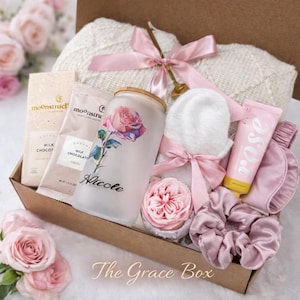 May include: A gift box filled with self-care items. Includes a white blanket, a glass with a rose design, chocolate, a pink eye mask, a tube of rose-scented lotion, a pink satin scrunchie, and a decorative rose. The box is tied with pink ribbons.